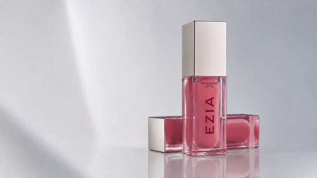 EZIA pH Adaptive Lip Oil in a clear glass bottle with a champagne cap, shade Silk Vanilla, against a soft draped backdrop